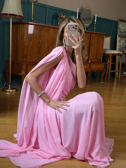 Woman in a pink saree sitting on the floor in a room with wooden furniture and a lamp.