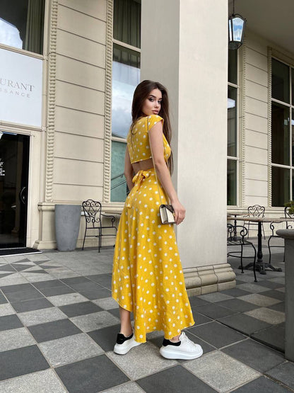 Woman in a yellow polka dot dress standing on a checkered pavement with a building in the background.