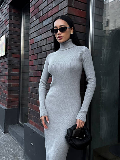 Woman in a gray turtleneck dress standing against a brick wall.