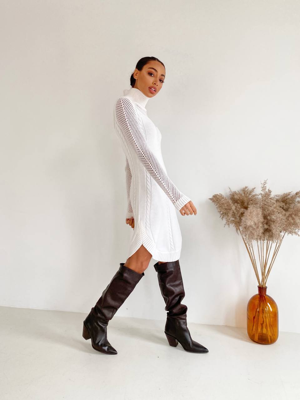 Woman in a white dress and brown knee-high boots standing against a plain background with a vase of dried plants.