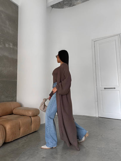Person wearing a long brown coat and blue jeans standing in a minimalistic room with a brown sofa and white walls.