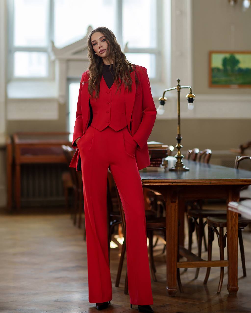 Woman in a red suit standing in a room with wooden furniture and large windows.