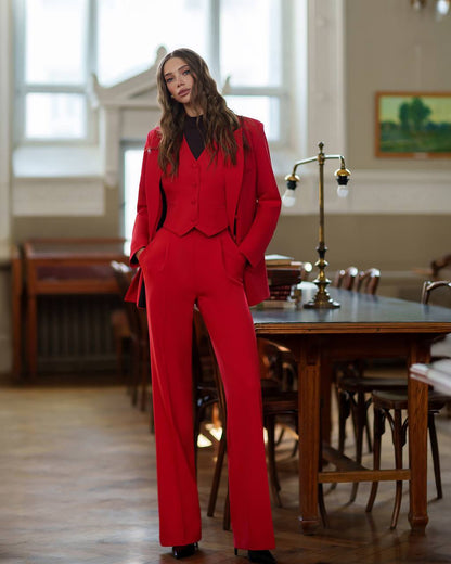 Woman in a red suit standing in a room with wooden furniture and large windows.