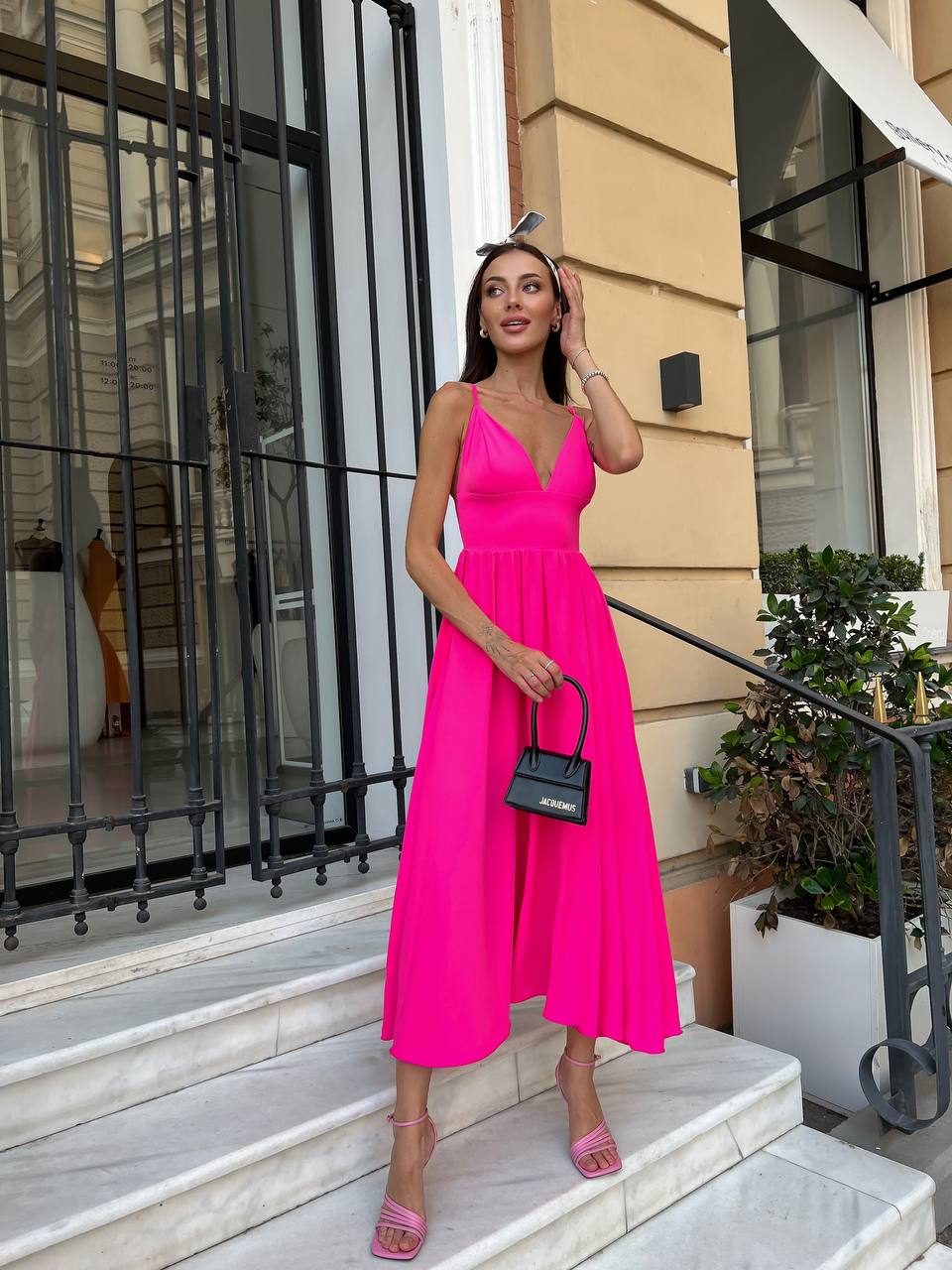 A woman stands on outdoor stone steps in front of a beige building, wearing a bright hot pink sleeveless midi dress with a deep V-neckline and flared skirt. She is holding a small black handbag and wearing pink high-heeled sandals.