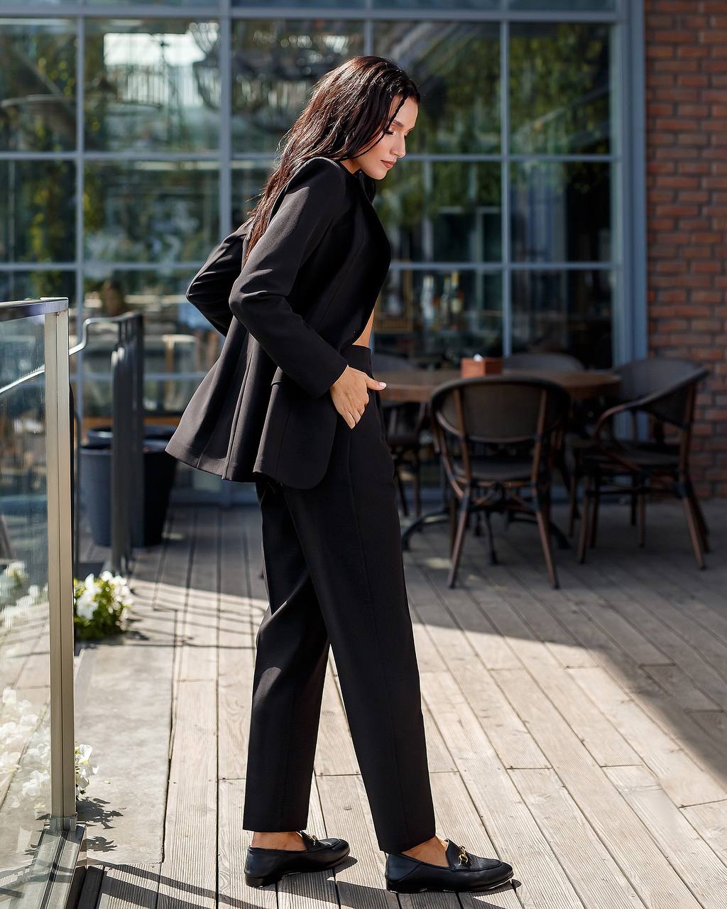 Woman in a black suit standing on a wooden deck with tables and chairs in the background.