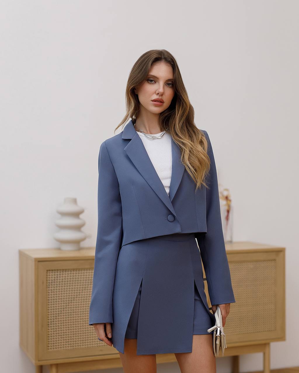 Woman wearing a blue suit standing in a room with a wooden cabinet and decorative items.