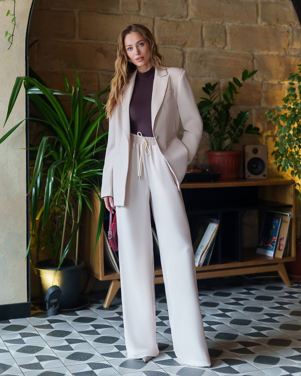 Woman in a beige suit standing in a room with plants and a fireplace.