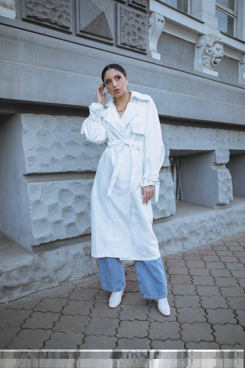 Woman in a white trench coat and blue pants standing in front of a stone building.
