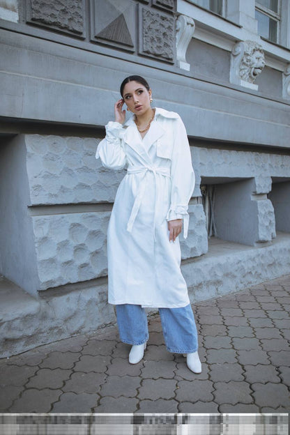 Woman in a white trench coat and blue pants standing in front of a stone building.