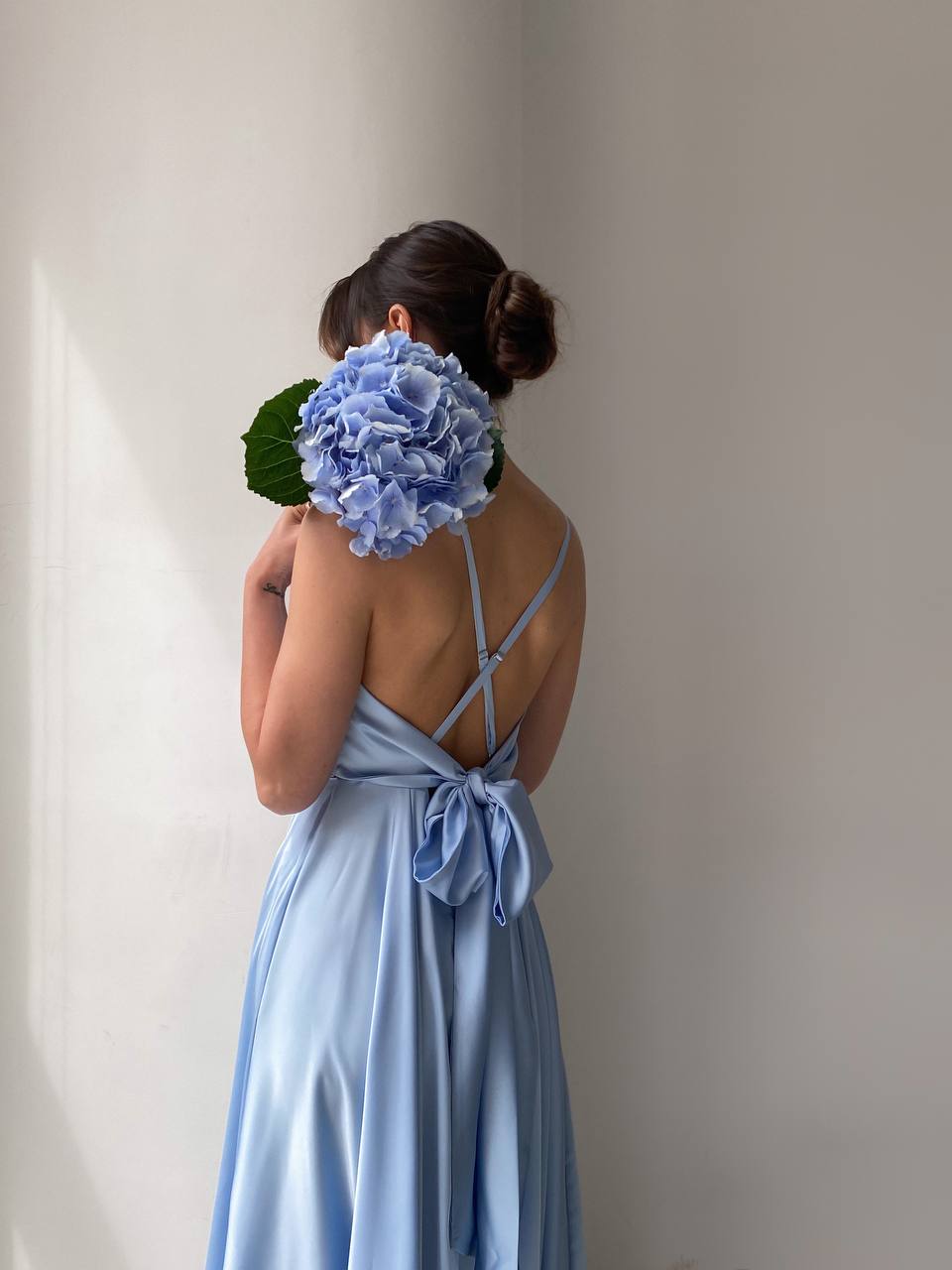 Woman in a light blue dress holding a large blue flower against a plain background
