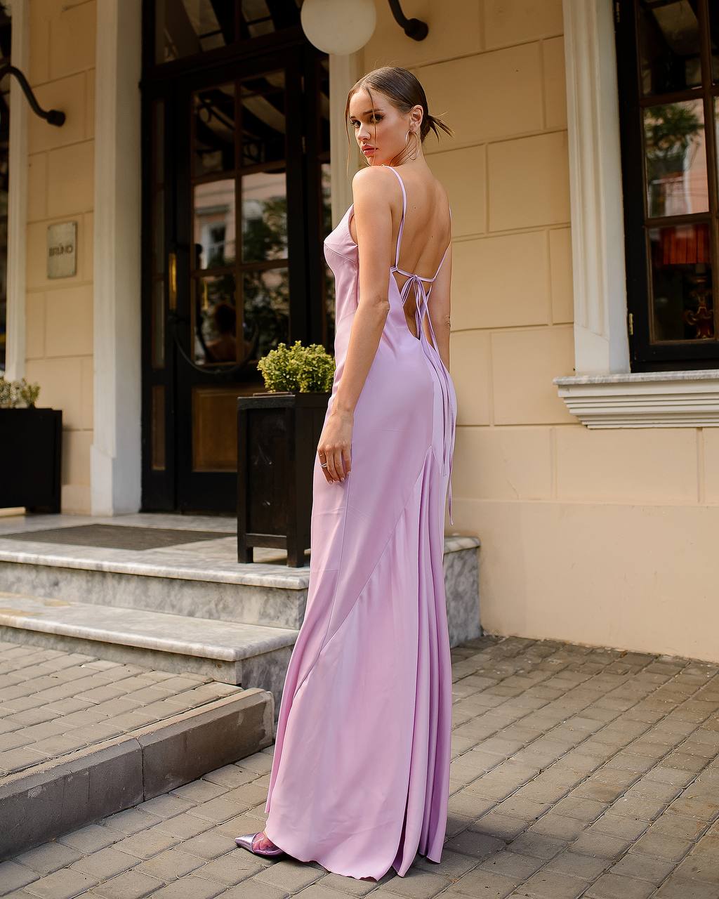 Woman in a long pink dress standing on a sidewalk in front of a building.