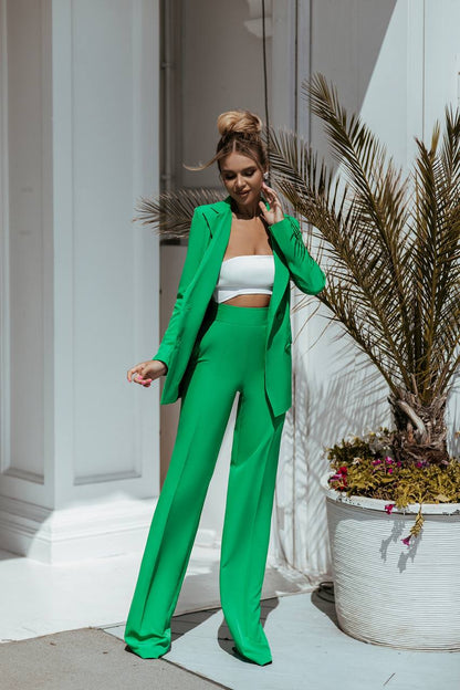 Woman in a green suit standing outdoors with plants and a building in the background