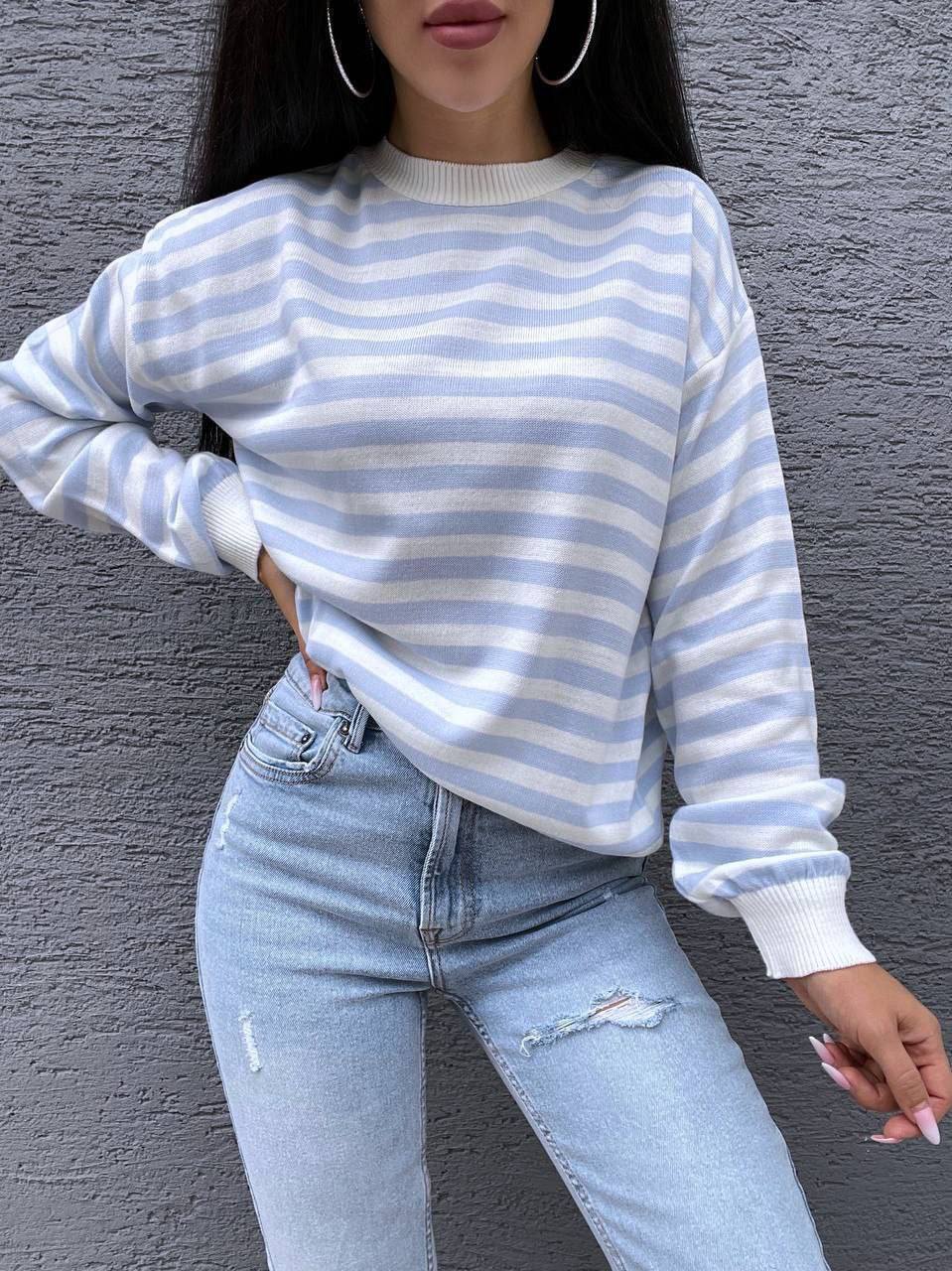 Person wearing a blue and white striped sweater and light blue jeans against a gray wall.