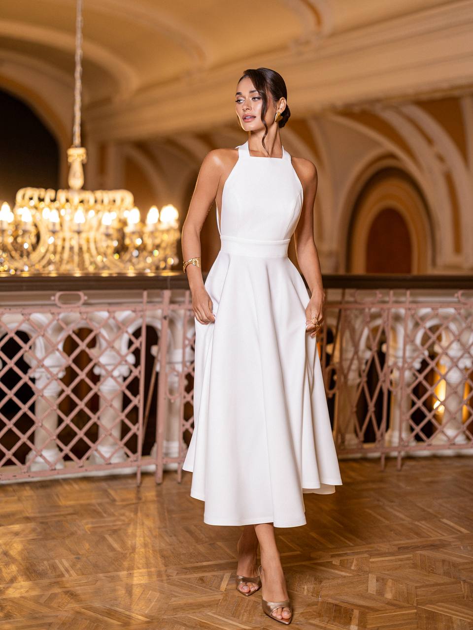 Woman in a white dress standing in an elegant indoor setting with chandeliers.