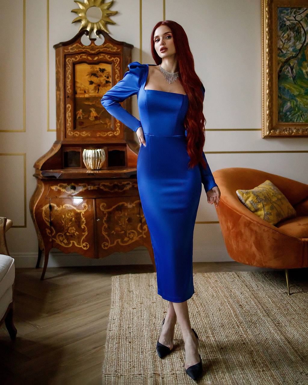 Woman in a blue dress standing in an elegant room with ornate furniture and a painting.