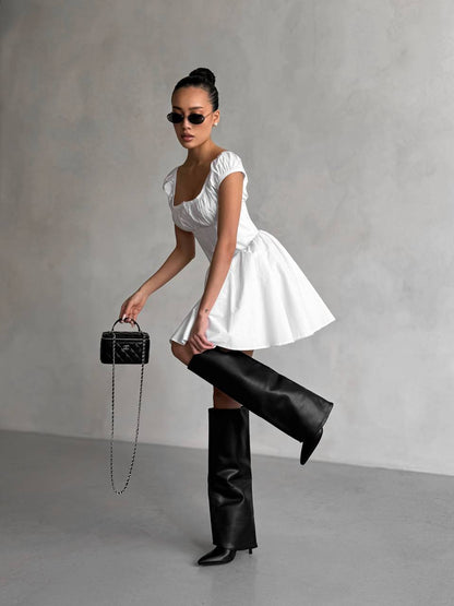 Woman in a white dress and black knee-high boots holding a black handbag against a gray background