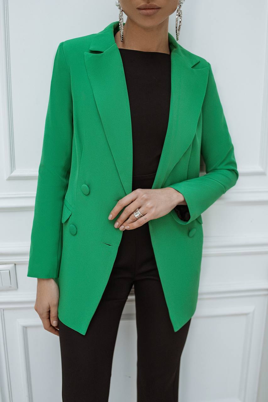 Person wearing a green blazer over a black top against a white paneled wall.