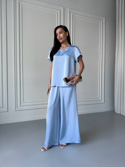 Woman wearing a light blue outfit standing against a white paneled wall.