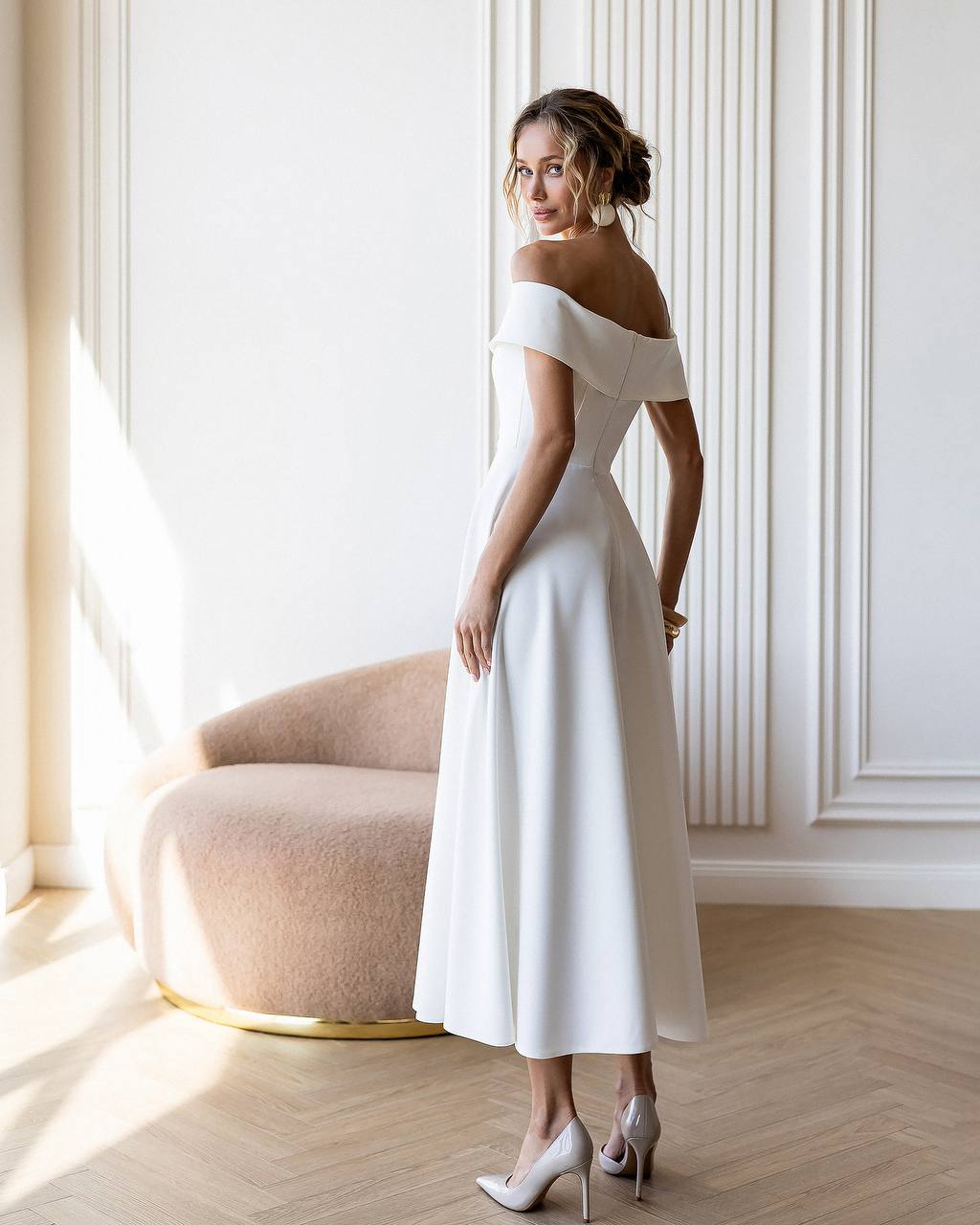 Woman in a white off-shoulder dress standing in a bright room with a chair and window.