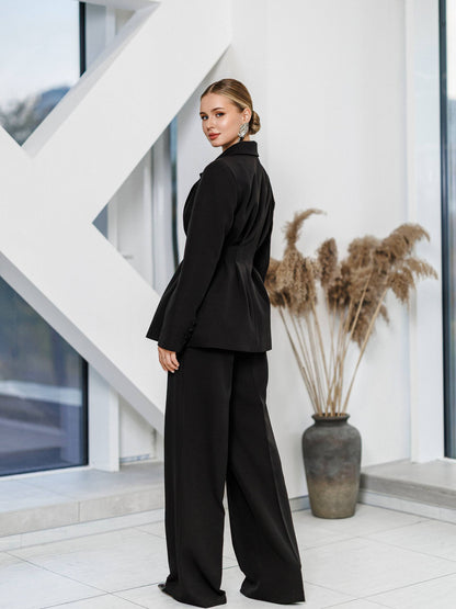 Woman in a black suit standing in a modern indoor setting with white walls and decorative plants.