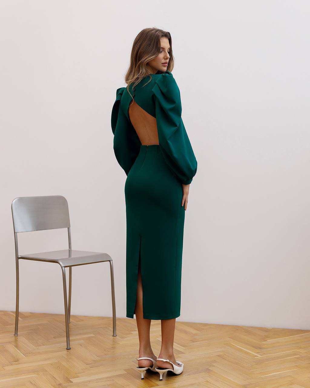 Woman wearing a green dress with an open back and slit skirt, standing in a room with a chair and wooden floor.