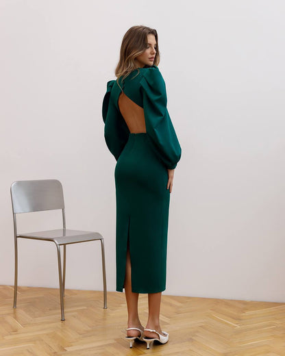 Woman wearing a green dress with an open back and slit skirt, standing in a room with a chair and wooden floor.