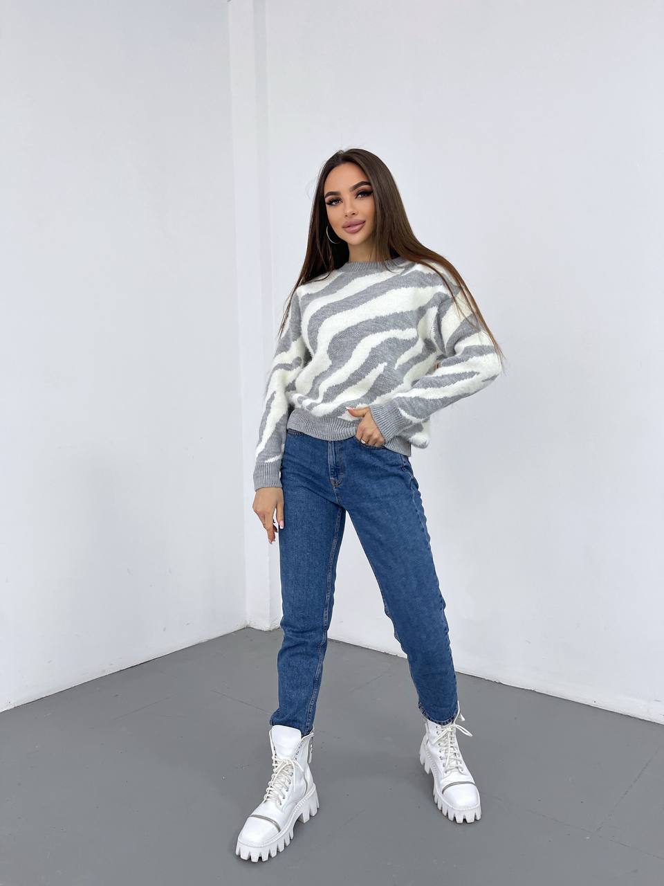 Woman wearing a striped sweater, blue jeans, and white boots in a minimalistic setting