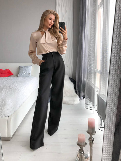 Woman taking a mirror selfie wearing a beige blouse and black high-waisted pants in a bedroom.