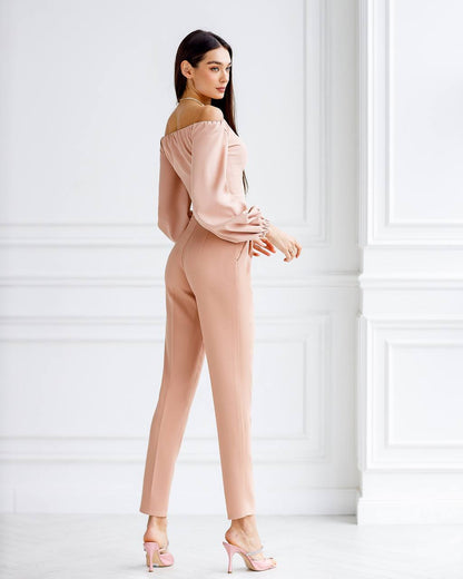 Woman wearing a beige off-shoulder top and pants against a white wall.