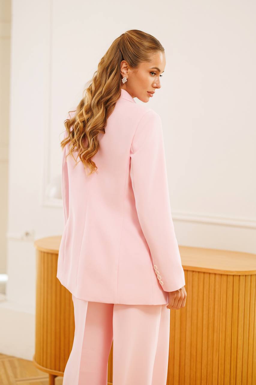 A woman with long wavy light brown hair is shown from the back wearing a light pink tailored blazer and matching wide-leg trousers in a bright indoor setting with a light wood cabinet.