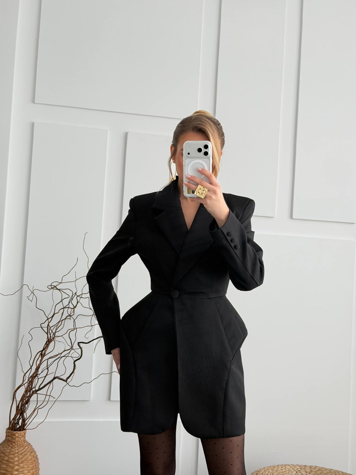 A woman takes a mirror selfie wearing a black belted blazer-style mini dress with structured shoulders and short skirt panels, paired with sheer black tights. She stands against a white paneled wall with a decorative dried branch arrangement in a woven vase on the left.