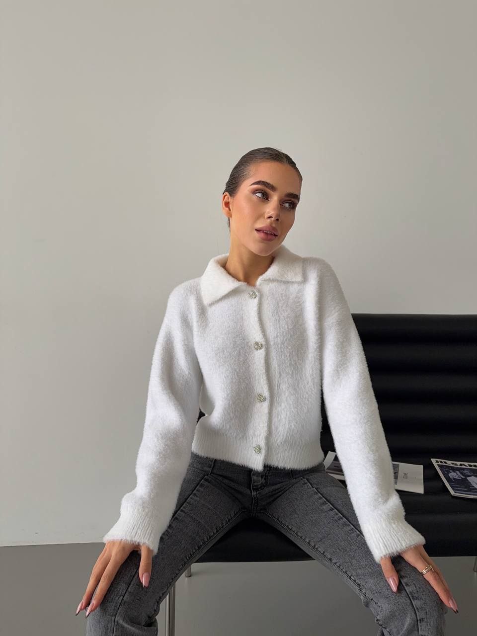 Woman wearing a white cardigan sitting on a black chair against a plain background