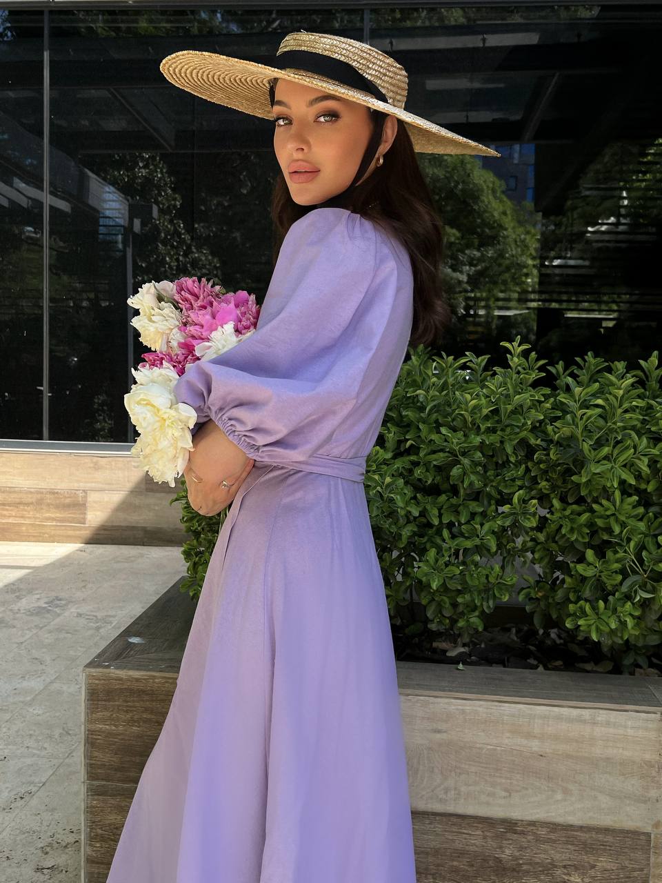 Woman in a lavender dress and straw hat holding flowers outdoors.