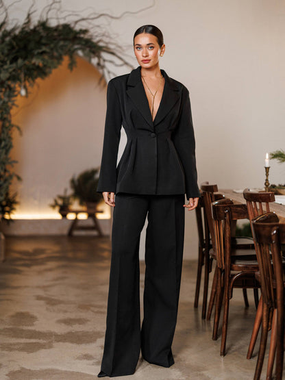 Woman in a black suit standing in a room with wooden furniture and plants.