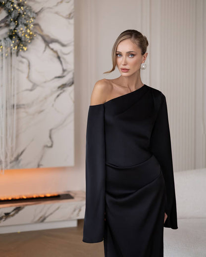 A woman wearing a black one-shoulder draped dress with long sleeves, standing in a modern interior with a marble wall and fireplace visible in the background.