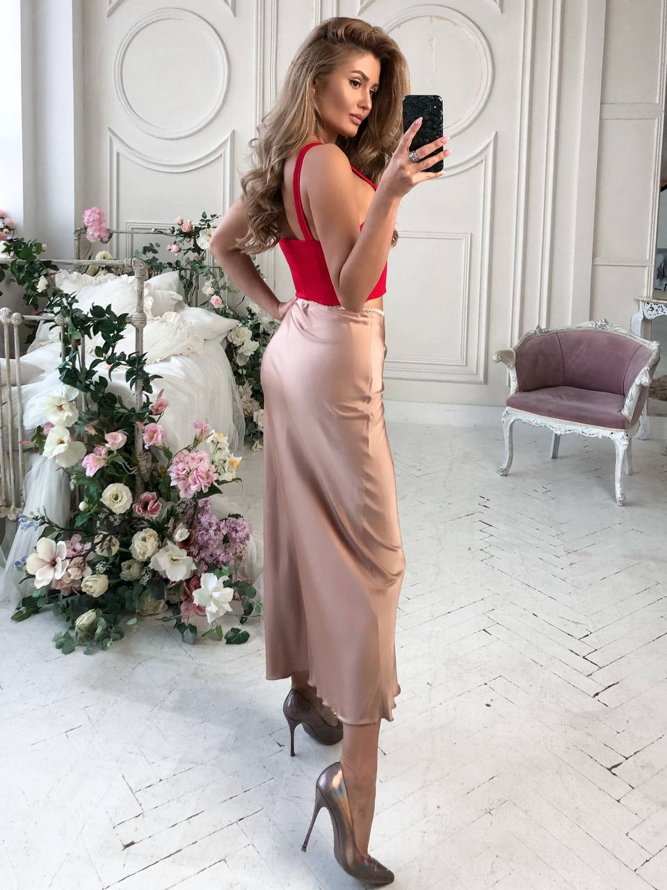 Woman taking a selfie in a red top and beige skirt in a decorated room.