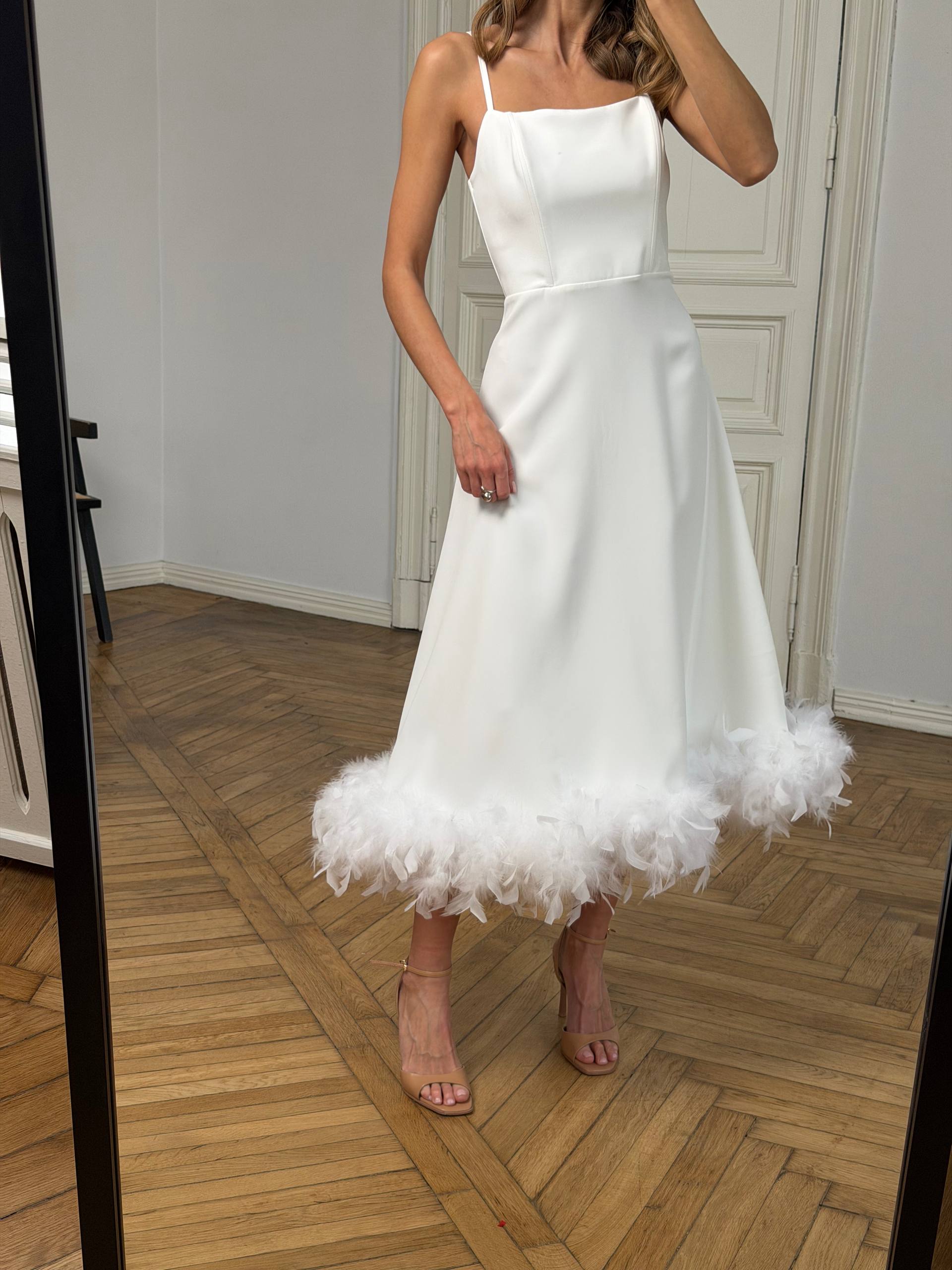 Woman wearing a white dress with a feathered hem in a room with wooden flooring.