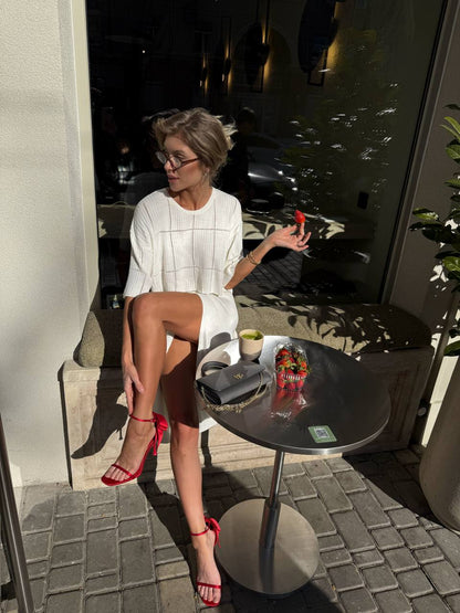 Woman sitting at a table outdoors, holding a phone, with a drink and a plant in the background.