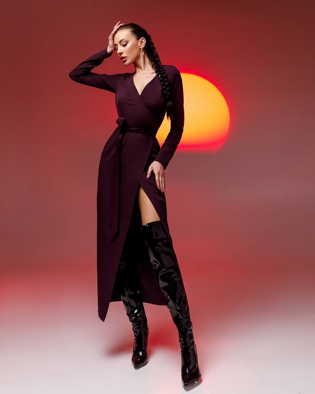 Woman in a long dark purple dress with a high slit, standing against a gradient background with warm tones.