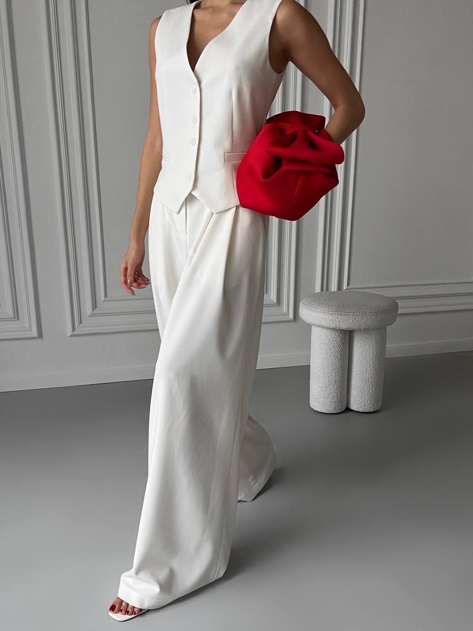 Person wearing a white outfit holding a red clutch against a gray wall.