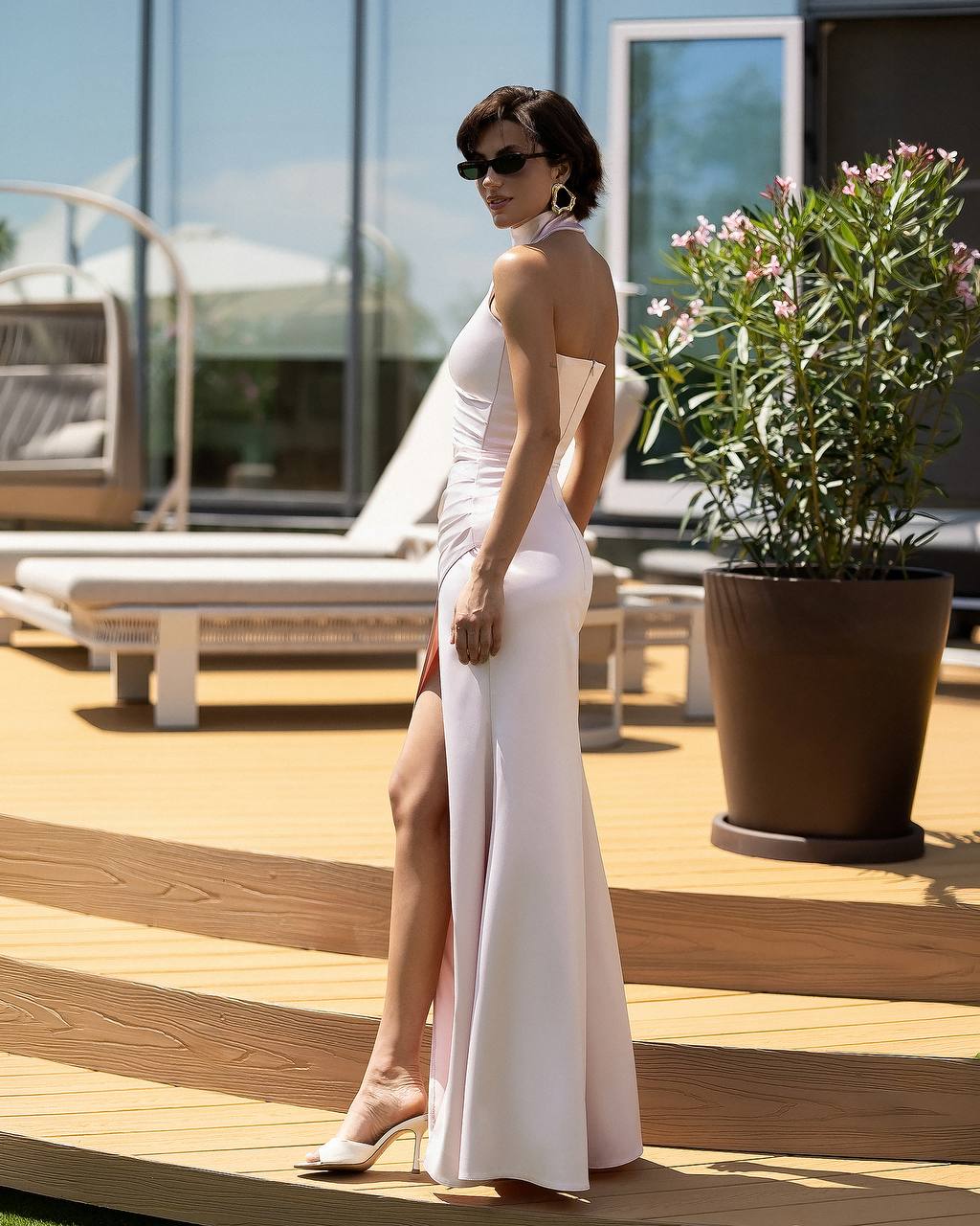 A woman wearing a sleeveless white halter-neck maxi dress with a thigh-high slit, styled with sunglasses and heels, standing on a wooden deck next to a potted plant.