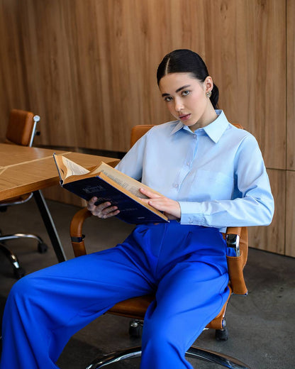 Woman in a light blue shirt and blue pants sitting on a chair, holding an open book.