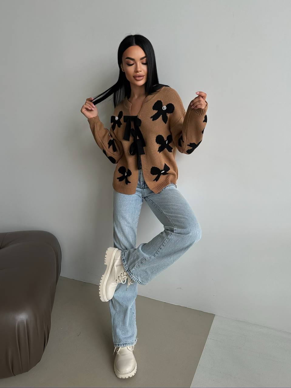 Woman wearing a brown sweater with black patterns, light blue jeans, and white sneakers in a minimalistic setting.