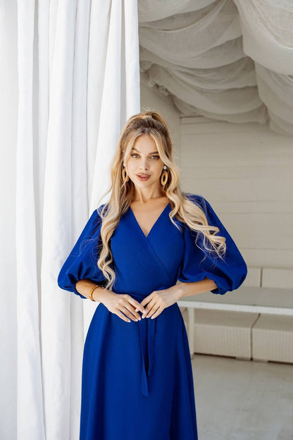 Woman in a blue dress standing in a room with white curtains.