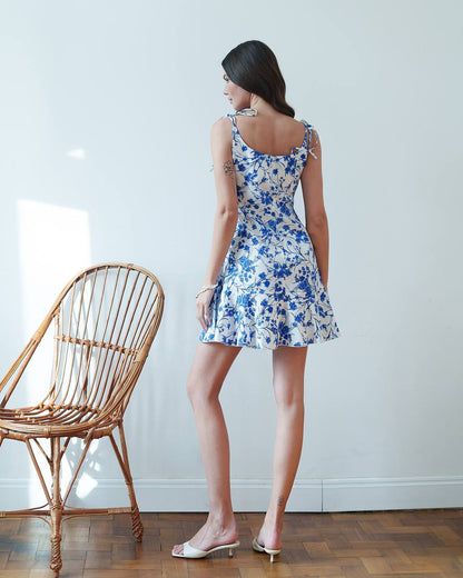 Woman wearing a blue floral dress standing next to a wicker chair indoors.