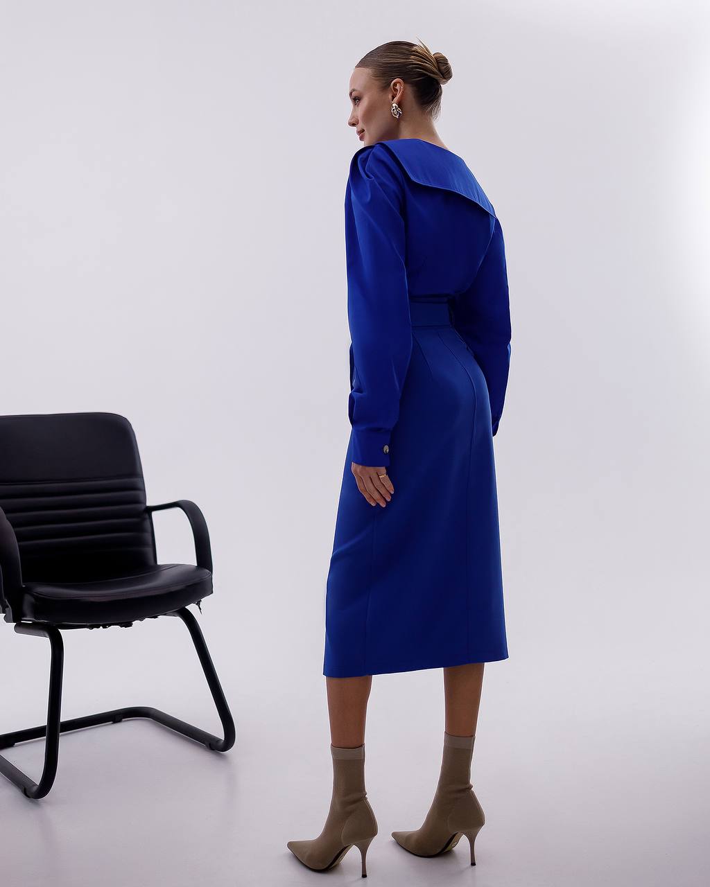 Woman wearing a blue dress standing next to a black chair on a white background