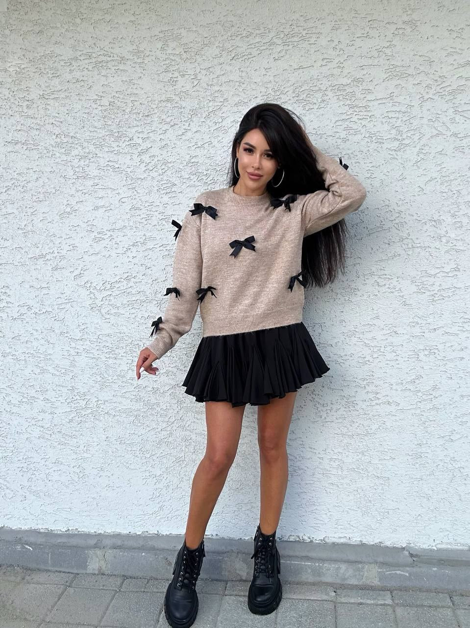 Woman wearing a beige sweater with black bow details and a black pleated skirt against a white wall.