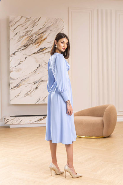 Woman in a light blue dress standing in a stylish room with a marble wall and beige armchair.