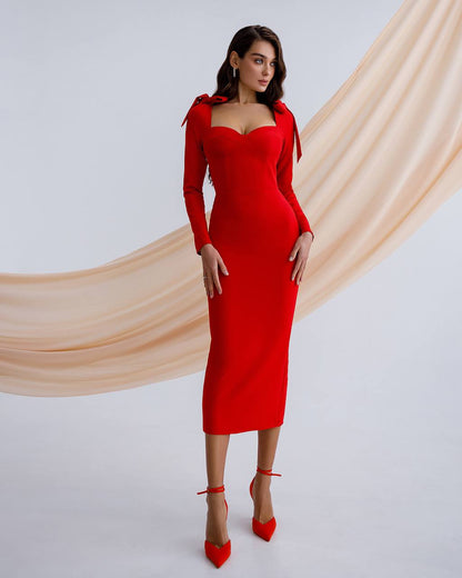 Woman wearing a red dress with long sleeves and a cutout design, standing against a beige curtain backdrop.