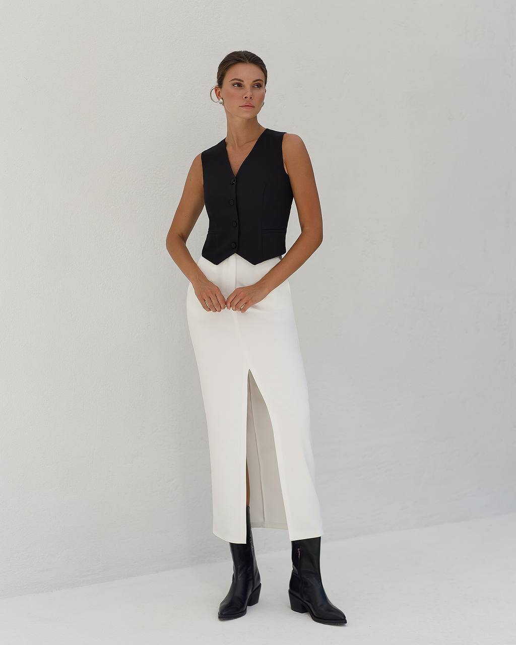 Woman wearing a black sleeveless top and white pants with a slit on a plain background